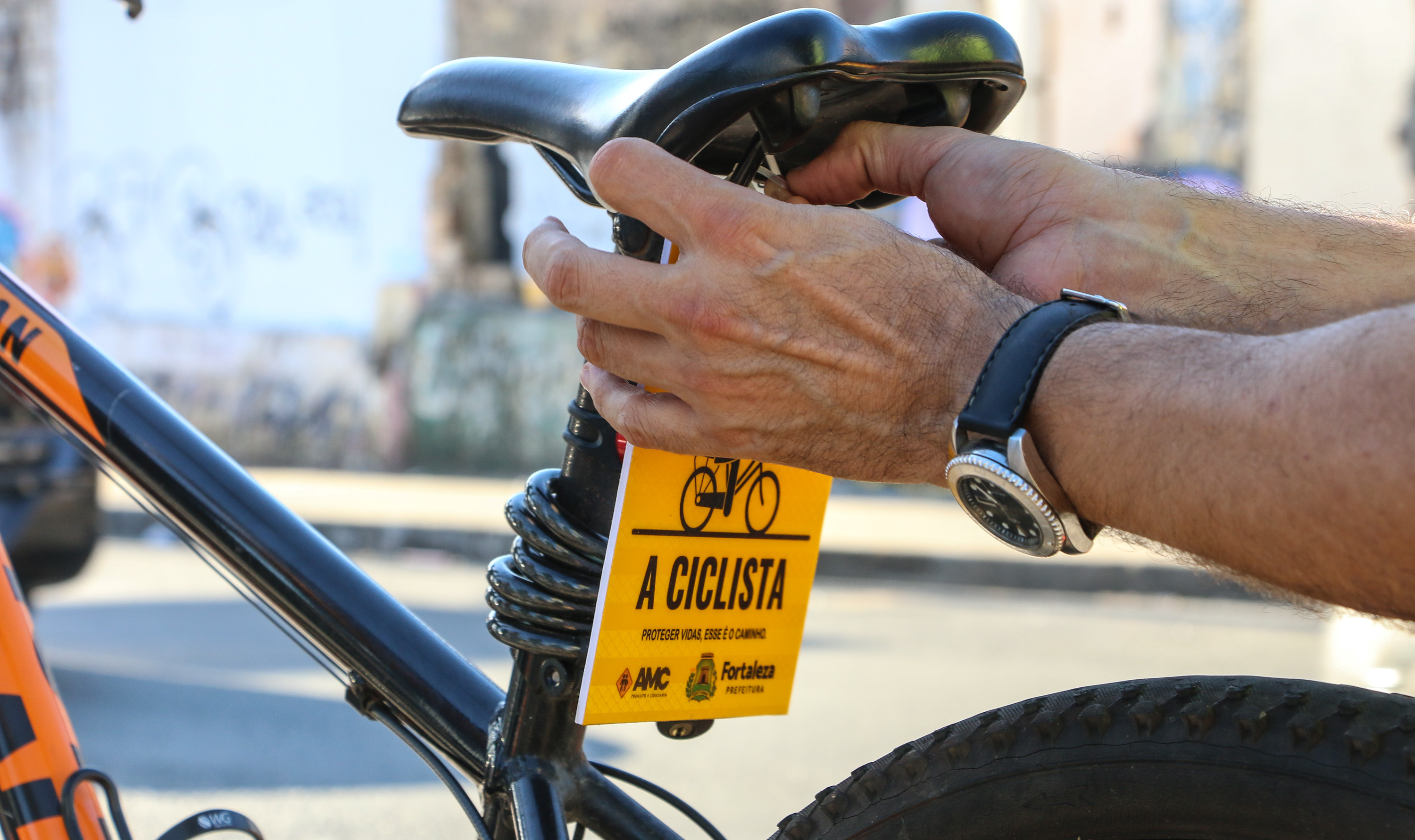 a foto mostra duas mãos colocando uma placa amarela com a frase respeite os ciclistas atrás do selim de uma bicicleta